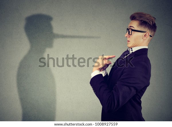 Side view of man in suit pointing at\
himself looking at shadow with long nose of a liar.\
