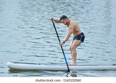 Side view of man training with sup board, roaming with long puddle in city lake. Male model wearing sport shorts, standing on big long board, swimming on water surface. Maintaing healthy lifestyle.