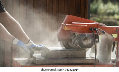 Side View Of A Man Using An Angle Grinder Or Circular Saw Outdoors To Cut Through A Block In A Cloud Of Dust.