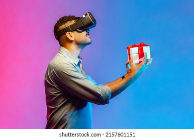 Side View Of Man In VR Virtual Reality Glasses Holding Wrapped Gift Box And Smiling Joyfully, Giving Present, Celebrating Holiday. Indoor Studio Shot Isolated On Colorful Neon Light Background.