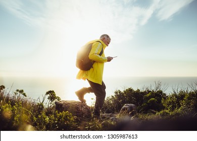 Side View Of A Man Wearing Backpack Walking On A Rocky Hill. Senior Man Hiking On A Hill Looking At His Mobile Phone With Sun In The Background.