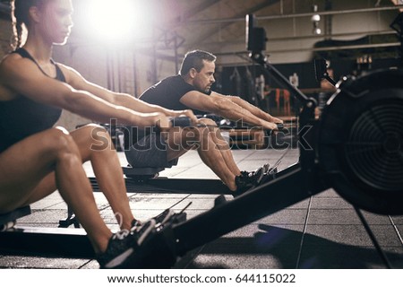 Side view of man and woman doing exercises with rowing machine at gym.
