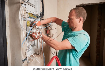 Side view of man in work gloves fixing electrical switchboard or distribution board in apartment under renovation. Electrician repairing household electrical panel with wires and switches.