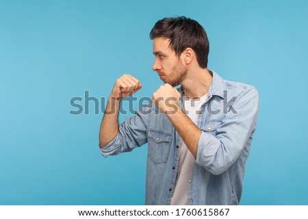 Side view of man in worker denim shirt standing with boxing gesture, punching to camera, provoking fight or ready to self-defense, struggle concept. indoor studio shot isolated on blue background