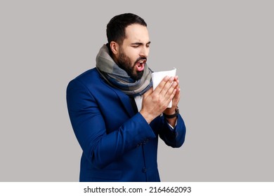 Side View Of Man Wrapped In Warm Scarf Sneezing In Napkin, Cleaning Runny Nose, Suffering Allergy Or Flu Symptoms, Seasonal Influenza Epidemic. Indoor Studio Shot Isolated On Gray Background.