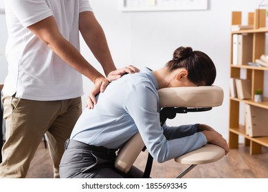 Side view of masseur doing seated massage of back for brunette businesswoman with blurred office on background