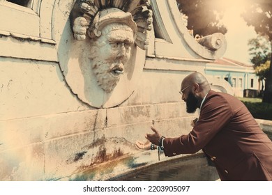 A Side View Of A Mature Bald Black Man With A Beard, In A Tailored Suit Of A Chestnut Color And Eyeglasses, Washing His Hands In The Water Of A Street Antique Marble Fountain Or Going To Drink From It