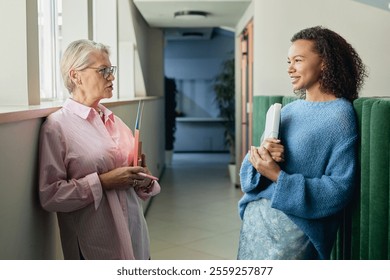 Side view of mature businesswoman casually chatting with younger employee of multiethnic background establishing intergenerational contact in workplace while standing in hallway, copy space - Powered by Shutterstock