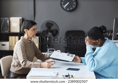 Side view of mature female manager passing rejected visa application form to anxious black woman touching her head while feeling upset