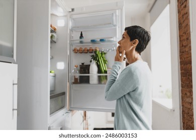 Side view of mature female with short hair standing in front of opened refrigerator at home with puzzled pensive facial expression, thinking of cooking breakfast, looking for ingredients