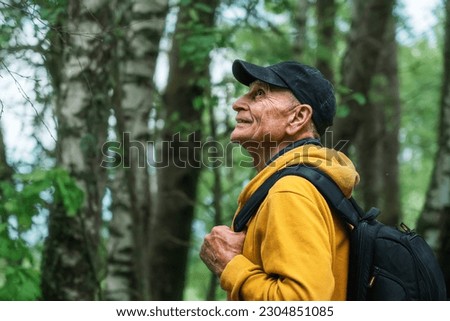Side view of mature hiker wearing cap and holding backpack standing in summer forest with birch trees. Tourism concept.