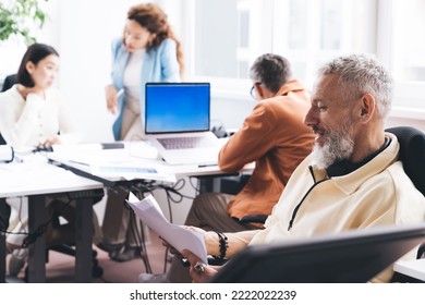 Side View Of Mature Male Entrepreneur Sitting On Chair And Reading Documents While Working On Business Project With Colleagues In Modern Office