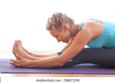 Side View Of A Mature Professional Woman Exercising And Stretching Her Body Doing Flexibility Exercises And Bending Her Back, Sitting On A Yoga Mat Indoors. Senior Woman Fit And Healthy Lifestyle.