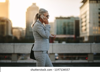 Side View Of Mature Woman In Business Suit Walking Back To Home Talking On Phone. Senior Businesswoman Using Cellphone On City Street With Urban Background.