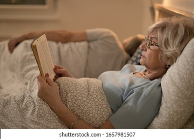 Side View Of Mature Woman Lying Down In Bed And Reading Book. Her Husband Is Sleeping In The Background. 