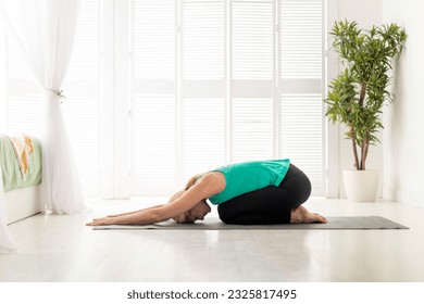 Side View Of Mature Woman Practicing Yoga, Meditating In Child's Pose With Face Down, Senior Female In Activewear Exercising On Fitness Mat Indoors, Sporty Lady Working Out In Light Room, Copy Space - Powered by Shutterstock