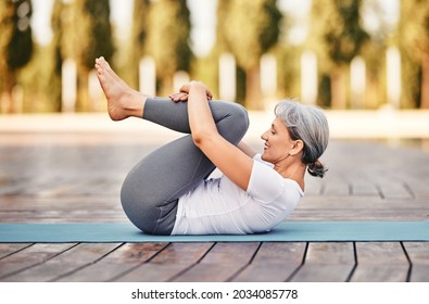 Side View Of Mature Woman In Sports Wear Lying On Mat Hugging Her Knees To Chest After Yoga Practice Outdoor In Park, Healthy Active Senior Female Doing Stretching Exercises In Morning On Fresh Air