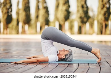 Side View Of Mature Woman In Sports Wear  Doing Plow Pose During Yoga Training In Park On Summer Day,  Healthy Active Senior Female Practicing Halasana On Mat In Morning On Fresh Air