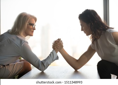 Side view mature and young businesswomen arm wrestling, confrontation concept, employees struggling for leadership at work, exerting pressure to each other, staring, sitting opposite in office - Shutterstock ID 1511348993