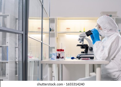 Side view of medical scientist in protective mask using microscope while studying dangerous coronavirus in laboratory