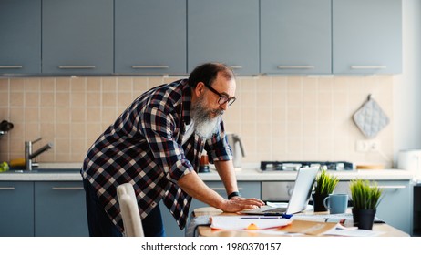 Side view medium shot of bearded senior man in checkered shirt and eyeglasses checking data on laptop computer bending over table in kitchen at home