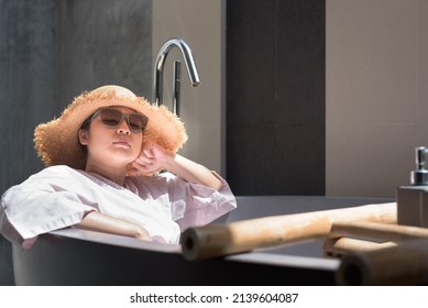 Side View, Medium Shot Portrait With Copy Space, Bored Young Adult Asian Woman Wearing Sunglasses, Hat, And Robe, Leaning Back In Outdoor Bathtub, Relaxing, Bathing Sun, Sleeping. Leisure Concept.