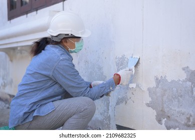 Side View, Medium Shot Of Senior Asian Woman Wearing Safety Hard Hat, Goggles, Gloves And Mask, Holding Trowel, Sitting, Peeling Off Paint Film From Old Wall. DIY Home Renovation Concept.