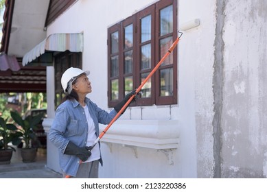 Side View Medium Shot Of A Senior Asian Woman Wearing White Safety Hard Hat, Gloves, Holding Paint Roller With Extension Handle, Painting Old House Wall. Active Retired Woman Fixing Home ,DIY Concept.