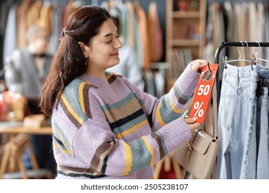 Side view medium shot of smiling Middle Eastern female customer choosing to buy second hand purse with fifty percent discount tag at thrift store