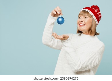 Side view merry smiling elderly woman 50s years old wear sweater red hat posing hold in hand toy bauble for Christmas tree isolated on plain blue background. Happy New Year celebration holiday concept