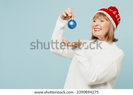 Side view merry smiling elderly woman 50s years old wear sweater red hat posing hold in hand toy bauble for Christmas tree isolated on plain blue background. Happy New Year celebration holiday concept