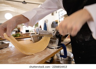 Side View Mid Section Of A Caucasian Senior Female Chef Wearing Chefs Whites And A Black Apron Preparing Fresh Pasta, Rolling Dough Through A Pasta Machine At A Cookery Class In A Restaurant Kitchen