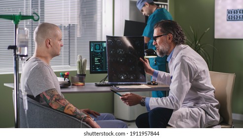Side View Of Middle Aged Doctor Demonstrating And Explaining CT Image To Male Oncology Patient While Sitting Near Computer During Chemotherapy In Hospital