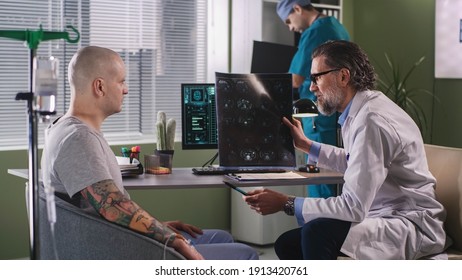 Side view of middle aged doctor demonstrating and explaining CT image to male oncology patient while sitting near computer during chemotherapy in hospital