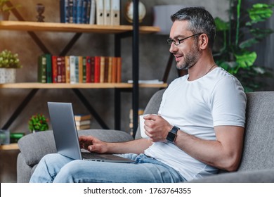 Side view of middle aged man drinking coffee while working on his laptop in living room