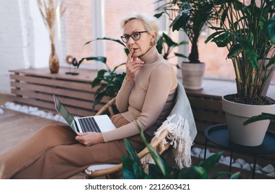 Side View Of Middle Aged Woman In Eyeglasses Sitting On Chair And Touching Lips While Watching Online Webinar Using Netbook In Living Room At Home