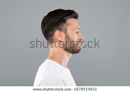 Side view of middle-aged bearded man in white t-shirt over grey studio background, copy space. Profile portrait of handsome man with closed eyes posing on gray, standing straight and looking aside