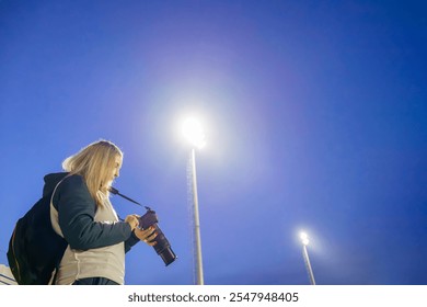 Side View of a Middle-Aged Female Sports Photographer Reviewing Photos on Her Camera at a Soccer Field During Twilight, with Stadium Lights and Sky in the Background - Powered by Shutterstock