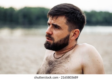 Side View Of Middle-aged Handsome Frowning Bearded Man With Short Dark Hair Sitting Shirtless On Beach Shore Bank Coast Of River Lake, Relaxing, Resting. Holiday, Weekend, Vacation, Summer Activities.