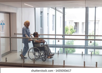 Side View Of Mixed-race Female Doctor In Hijab Pushing Senior Mixed-race Male Patient In Wheelchair At Corridor In Hospital.