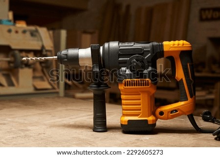 side view of a modern electric rotary hammer with vertical motor, mounted drill and additional handle. against the backdrop of a woodworking workshop