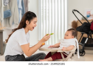 Side View Mother Feeding Her Asian Baby Daughter With Pumpkin Mashed Or Vegetable Mash On Rubber Spoon.Mom Trying To Feed Little Baby Boy At Home Enjoy And Spending Time Together.Baby Feeding Concept