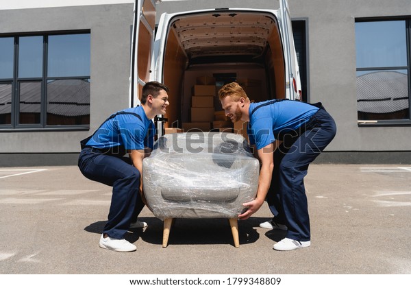 Side view of movers holding\
armchair in stretch wrap near truck with open doors on urban\
street