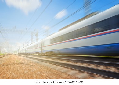 Side View Of The Moving Ultra High Speed Train Runs On Rail Way With Railway Infrustructure In The Blurred Background With Flare Effect. Electric High Speed Passenger Commuter Train. Intercity Express