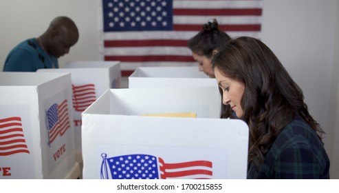 Side view MS of three people, two women and one man, Caucasian, Hispanic and African American, voting in booths at polling station. Large US flag on wall behind