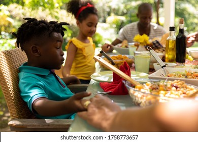 Side View Of A Multi-ethnic, Multi-generation Family Sitting Down At A Table Holding Hands With Eyes Closed And Saying A Prayer Before Having A Meal Together Outside On A Patio In The Sun.