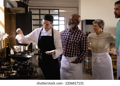 Side View Of Multi-ethnic Senior Group At Cookery Class, Discussing, Listening To A Teacher, Who Is Pouring Salt At A Frying Pan