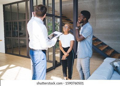 Side view of multiethnic smart business people with gadgets talking and discussing working process while man showing on glass wall in large spacious room 