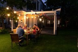 Side View Of A Multi-generation Caucasian Family Sitting In The Garden Outside Their House At A Dinner Table In The Evening For A Celebration Meal Together, Talking And Eating