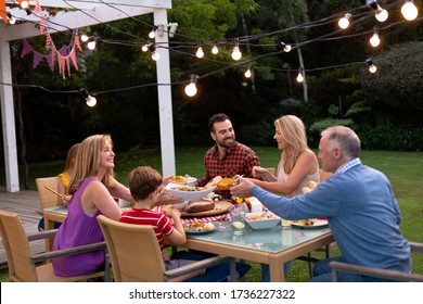 Side View Of A Multi-generation Caucasian Family Outside At A Dinner Table Set For A Meal, Sitting, Talking And Serving Food To Each Other